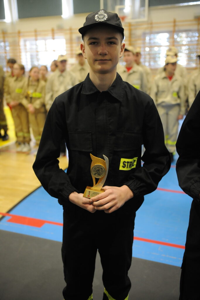 Młody chłopak w ciemnym mundurze strażackim trzyma złotą statuetkę. W tle widać grupę osób w beżowych mundurach. Scena dzieje się w jasnej sali gimnastycznej.