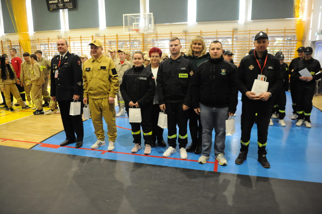 Grupa osób w strojach strażackich stoi na boisku sportowym. Tło to drewniane drabinki na ścianie i kosz do koszykówki. Dominują kolory żółty, czarny i niebieski.