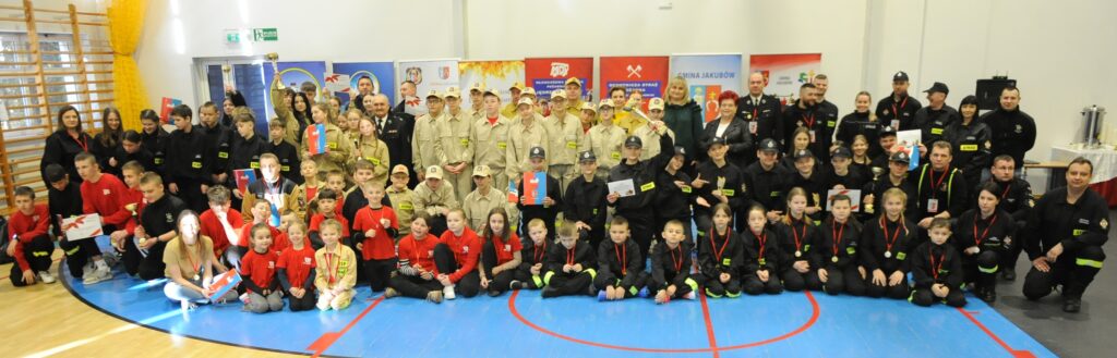 Grupa osób w strojach strażackich pozuje w sali z drewnianymi drabinkami. Tło to kolorowe bannery i biała ściana. Wszyscy mają medale lub dyplomy.