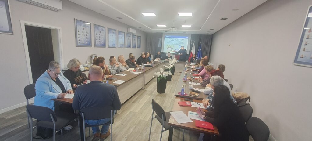 Na zdjęciu jest sala konferencyjna, ludzie siedzą przy długich stołach. Z przodu są kwiaty, w tle ekran z prezentacją. Ściany zdobią liczne plakaty.