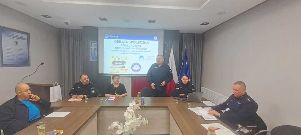 Na zdjęciu widoczni są funkcjonariusze policji prowadzący debatę o bezpieczeństwie seniorów. Uczestnicy siedzą przy stole z materiałami, widać flagi i projektor. Kolory dominujące to odcienie szarości i niebieskiego.