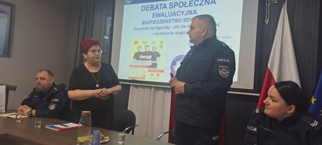 Na zdjęciu policjanci i kobieta prowadzą debatę o bezpieczeństwie seniorów. W tle widać prezentację i flagi Polski oraz UE. Sala jest w neutralnych kolorach z ciemnym tłem.