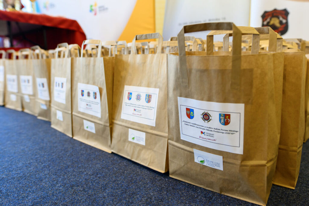 Papierowe torby stoją rzędem na niebieskim dywanie. Każda ma etykietę z herbem. Tło jest jasne, z kolorowymi elementami.