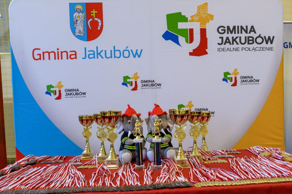 Na zdjęciu widać stoisko z pucharami na tle banera Gmina Jakubów. Są dwie maskotki słoni i czerwono-białe medale. Tło jest białe z kolorowym logo gminy.