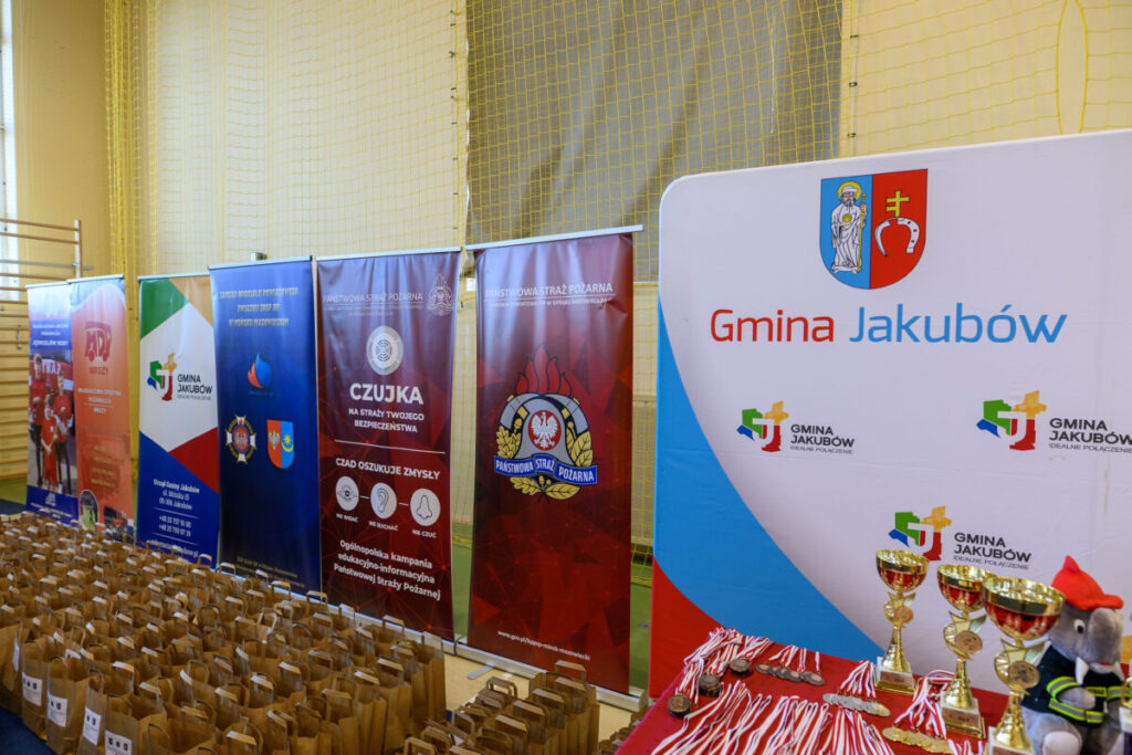 Bannery w jasnych kolorach promują Gminę Jakubów oraz straż pożarną. Na stole leżą medale, puchary i maskotka. Tło stanowi siatka w hali sportowej.