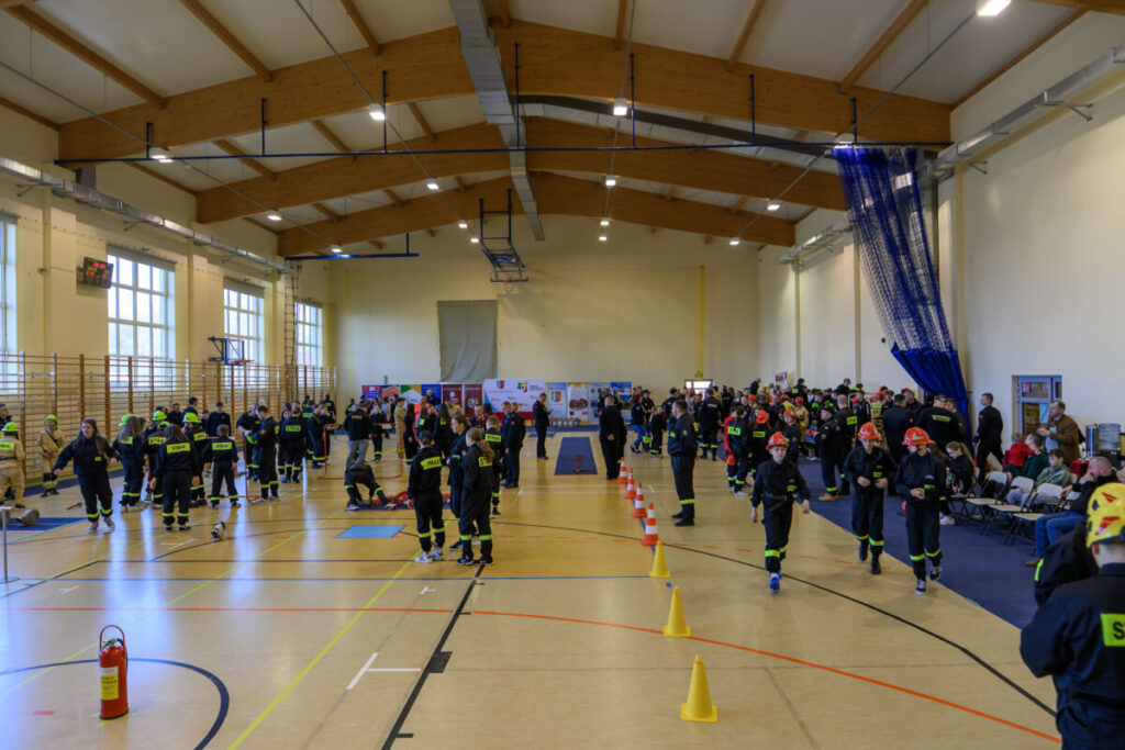 Dużo osób w strojach strażackich uczestniczy w zawodach w hali sportowej. Jasne ściany, drewniany sufit, liczne żółte pachołki na podłodze. Po prawej stronie siedzą widzowie.
