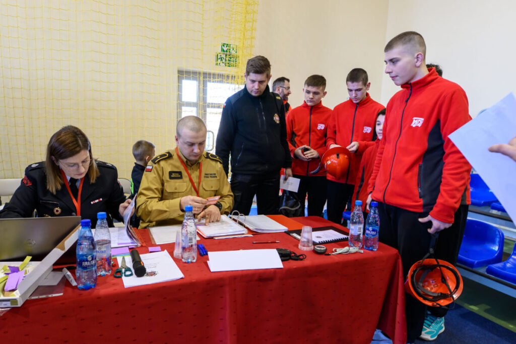 Grupa osób przy stole z czerwonym obrusem, w tym kobieta i mężczyzna w mundurach. Mężczyźni w czerwonych strojach stoją obok. Tło stanowi żółta siatka i ściana.