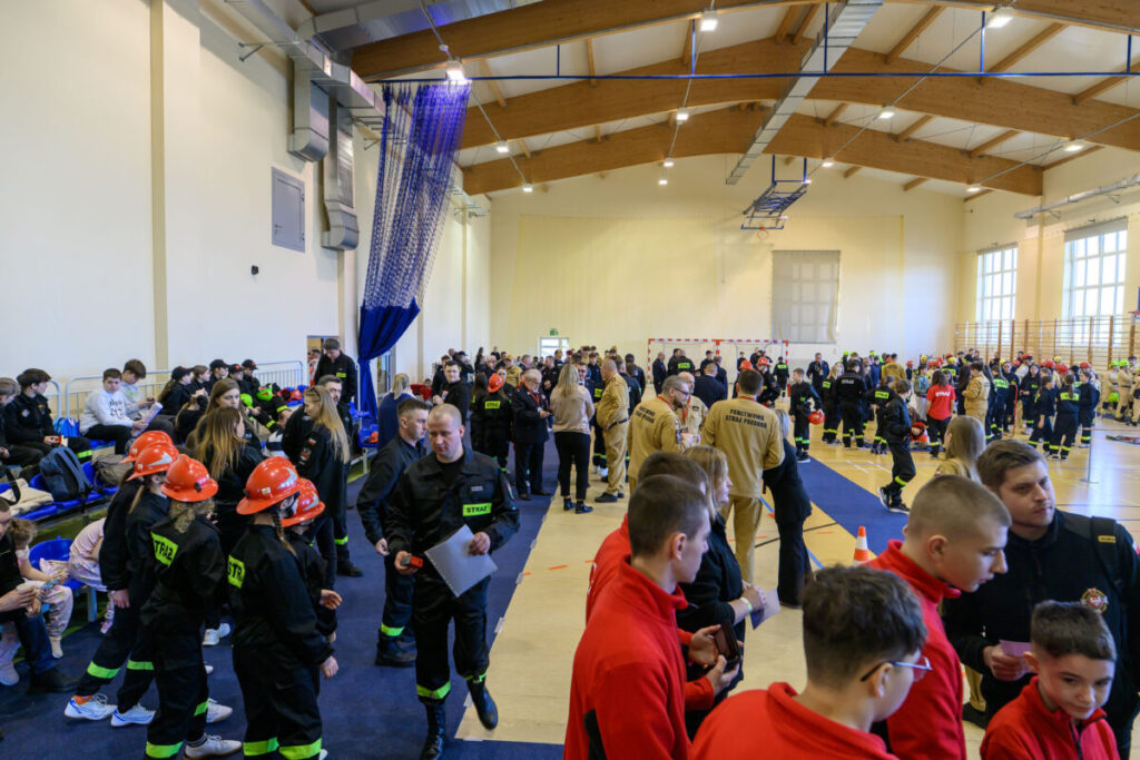 Tłum ludzi w strażackich mundurach w przestronnej sali gimnastycznej. Dominują kolory czarny i czerwony. W tle widać boisko i dużą siatkę.