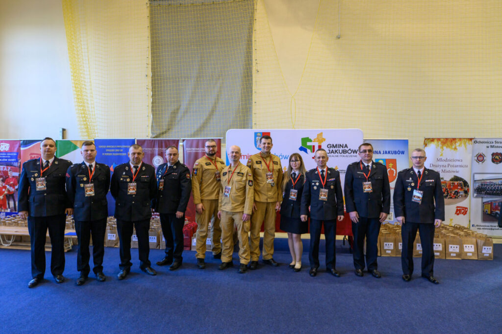 Grupa osób w mundurach strażackich na niebieskim dywanie, z banerami w tle. Dominują kolory czerwony, niebieski i żółty. Tło z siatką i logami gminy.