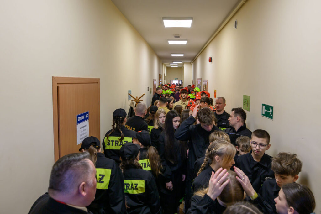 Grupa ludzi w czarnych mundurach strażackich stoi w wąskim jasnym korytarzu. Na drzwiach napis Szatnia WYC. W tle widać osoby w kolorowych kaskach strażackich.