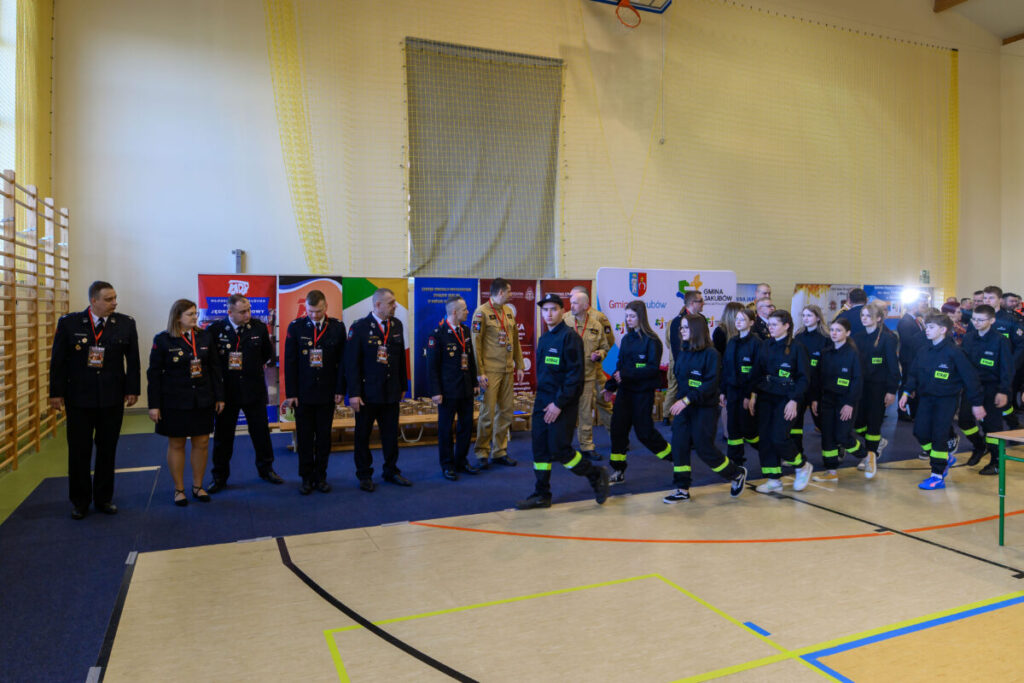 Grupa strażaków w czarnych mundurach stoi w sali gimnastycznej. Tło z kolorowymi banerami, jasne ściany. Obok maszeruje młodzież w podobnych uniformach.