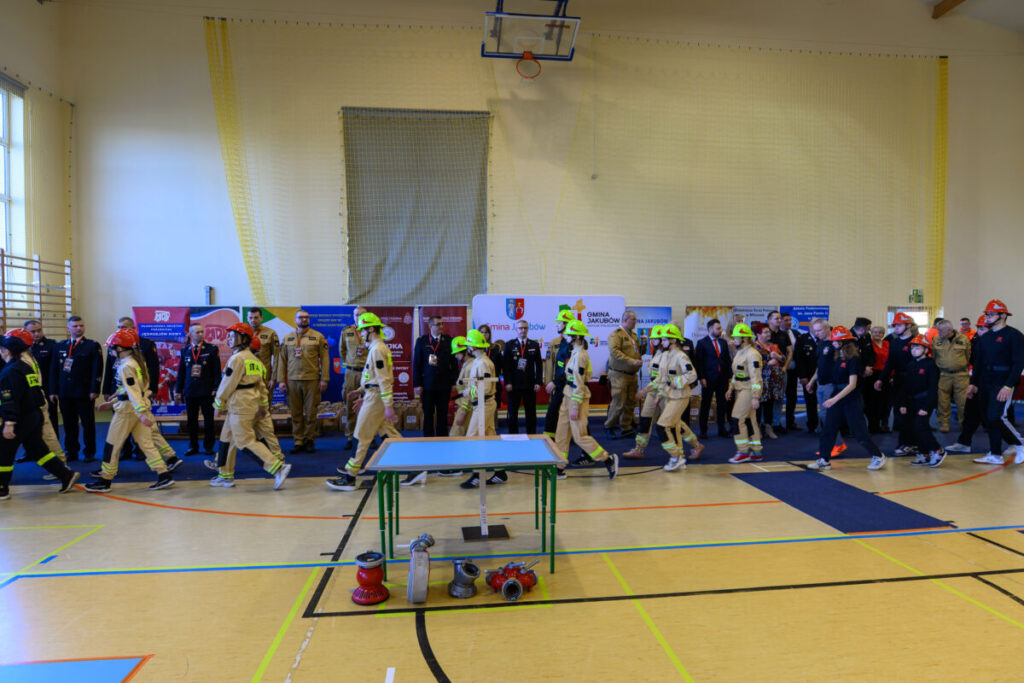 Grupa strażaków w hełmach w hali sportowej. Tło to kolorowe banery i żółta ściana. Na podłodze sprzęt strażacki.