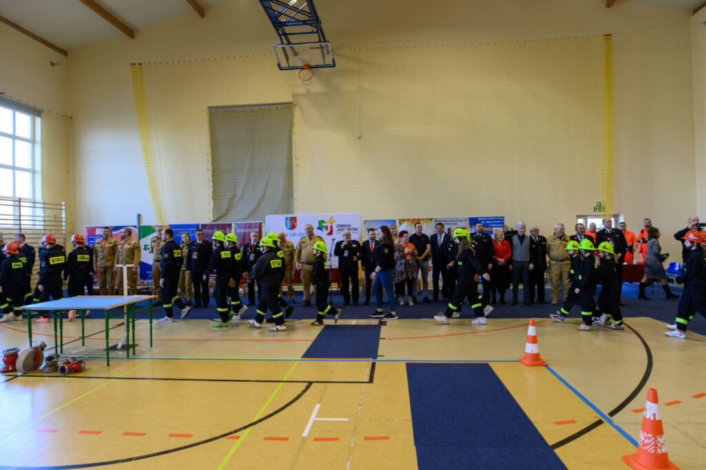 Grupa młodych strażaków w kaskach ćwiczy w hali sportowej. W tle stoją dorośli i plakaty. Podłoga jest żółta z niebieskimi elementami.