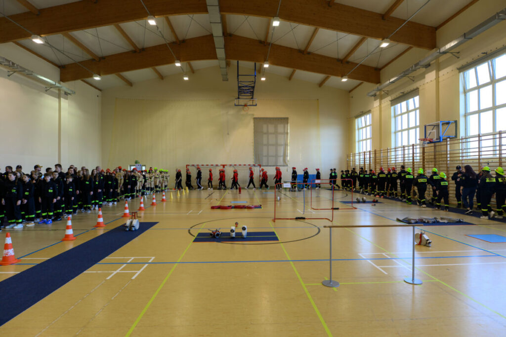 Ludzie w strojach strażackich stoją w sali gimnastycznej. Podłoga jest jasnobrązowa, wokół są pomarańczowe pachołki. Jasne ściany i duże okna w tle.