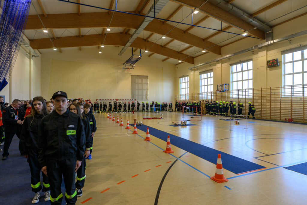 Grupa młodych strażaków w czarnych mundurach stoi w hali sportowej z drewnianym stropem. Na podłodze są rozstawione pachołki. Tło wypełniają jasne ściany z dużymi oknami.