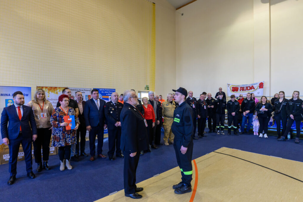 Grupa strażaków i urzędników na uroczystości w hali sportowej. Dominują ciemne stroje, niebieski dywan i jasne ściany. W tle bannery z napisem „RUDA”.