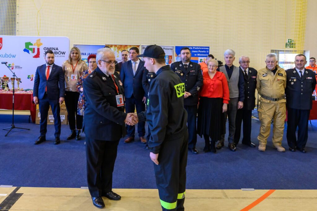 Grupa osób w odświętnych uniformach stoi w hali sportowej. Na pierwszym planie dwóch mężczyzn ściska sobie dłonie. Tło wypełniają kolorowe banery i krzesła.