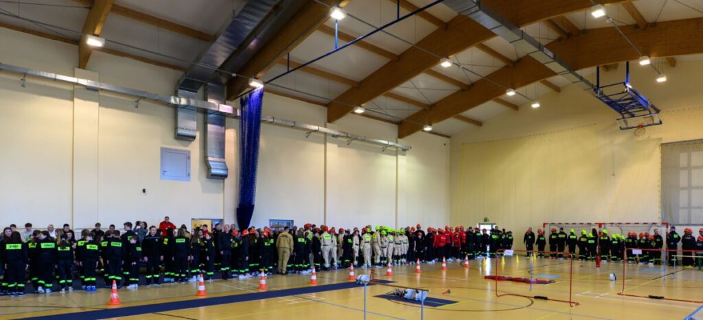 Grupa strażaków w różnokolorowych mundurach stoi na tle jasnej sali gimnastycznej. Na podłodze widoczne są pomarańczowe pachołki i sprzęt. Drewniany sufit i wentylacja podkreślają wnętrze.