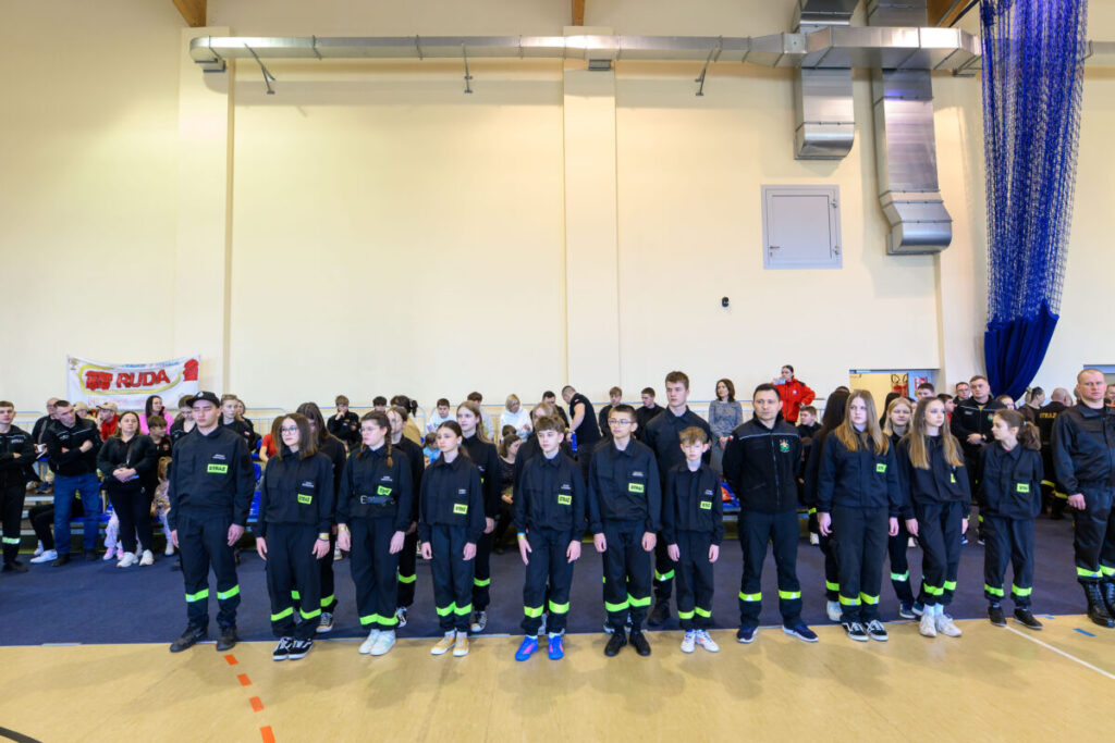 Grupa dzieci i młodzieży w ciemnych kombinezonach z zielonymi detalami stoi w jasnej sali. W tle siedzący widzowie i banner. Scena odbywa się w zamkniętym pomieszczeniu z jasnymi ścianami.