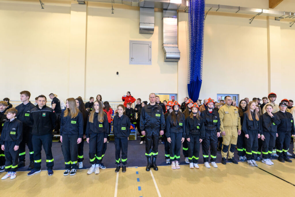 Młodzi ludzie w czarnych mundurach strażackich z zielonymi napisami stoją w hali sportowej. Na głowach mają czerwone kaski. Tło jasne, z widocznymi elementami wentylacji.