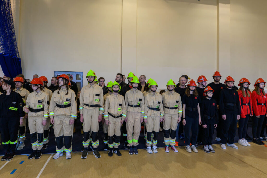 Grupa osób w strojach strażackich, kaski w kolorze zielonym i pomarańczowym. Tło beżowe, budynek wewnątrz. Główny kolor mundurów kremowy.