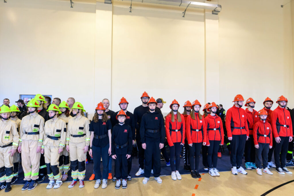 Grupa młodych osób stoi w dwóch rzędach, ubrana w stroje strażackie. Po lewej są beżowe mundury i zielone kaski, po prawej czerwone bluzy i pomarańczowe kaski. Tło jest jasne, wnętrze hali.
