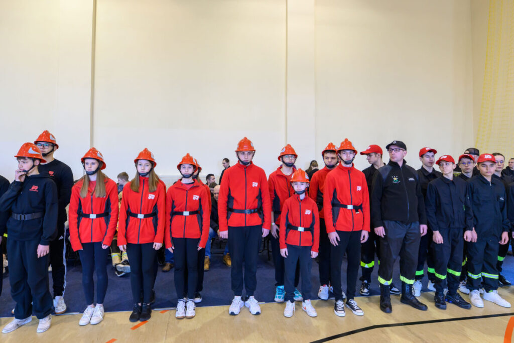 Grupa młodych ludzi w czerwonych kurtkach i pomarańczowych kaskach stoi na tle jasnej ściany. Część z nich nosi czarne stroje z zielonymi paskami. Pomieszczenie jest dobrze oświetlone.