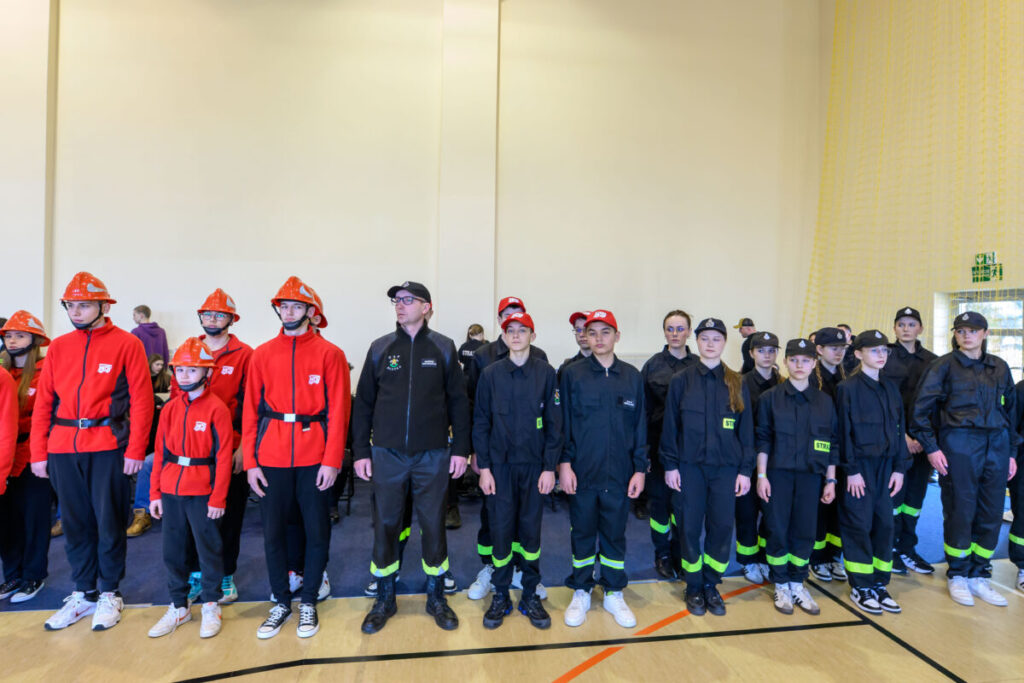 Grupa młodych ludzi w strojach strażackich; czerwone i czarne kurtki. Jasne wnętrze sali z żółtą siatką w tle.
