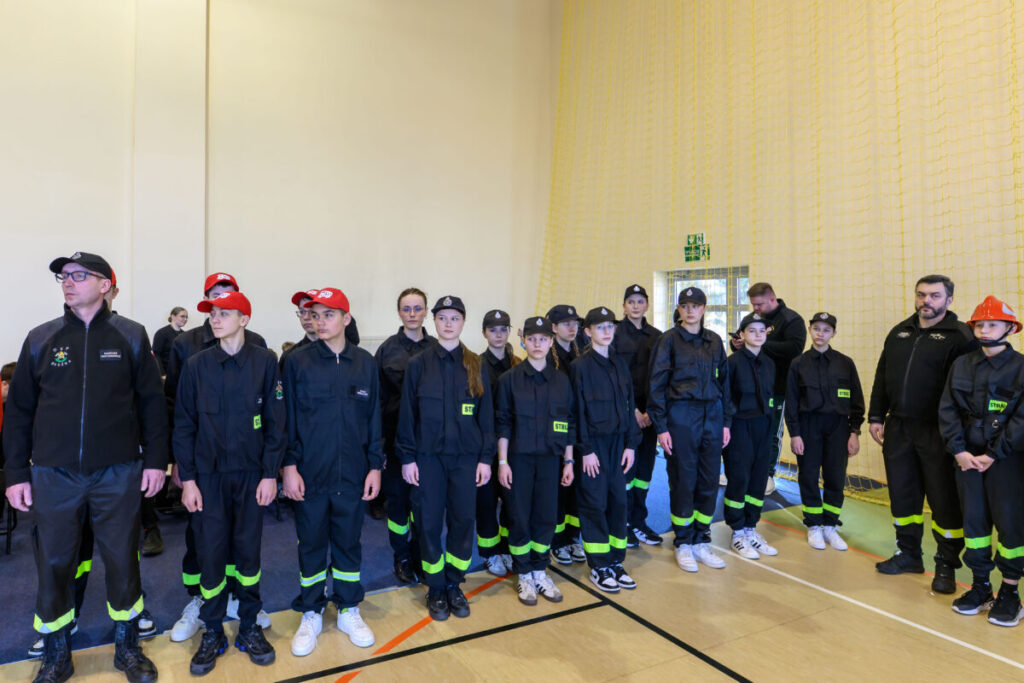 Grupa młodych osób w czarnych mundurach stoi w sali. Mają czapki i kolorowe akcenty na strojach. Tło jest jasne z żółtą siatką.