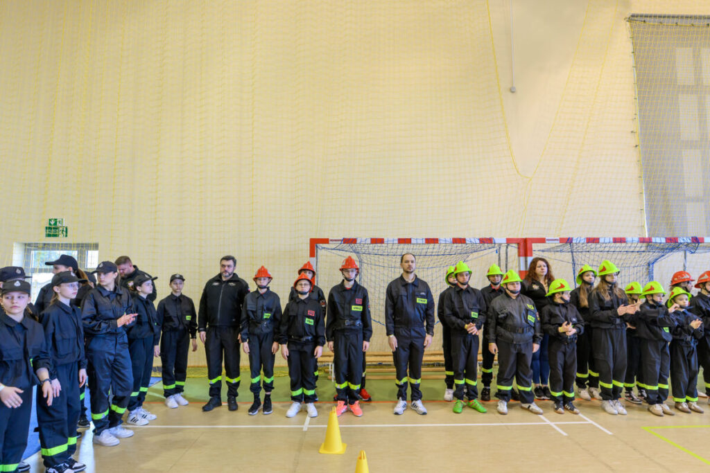 Grupa ludzi w strojach strażaków stoi w hali sportowej. Tło to siatka na sufit i bramka, dominują kolory czarny i czerwony. Na głowach zielone i czerwone hełmy strażackie.
