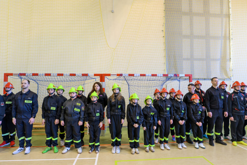 Grupa dzieci i dorosłych w stroju strażackim stoi na tle bramki w hali sportowej. Mają kaski w kolorze zielonym i czerwonym. Tło to żółta siatka na jasnej ścianie.
