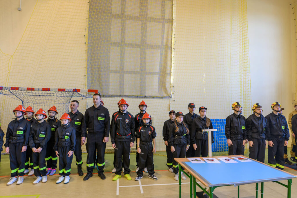 Grupa młodych strażaków w czarnych mundurach i czerwonych hełmach stoi na tle żółtej siatki w hali sportowej. Po lewej stronie znajduje się bramka, a po prawej stół z kartkami. Podłoga jest w kolorze jasnobrązowym,