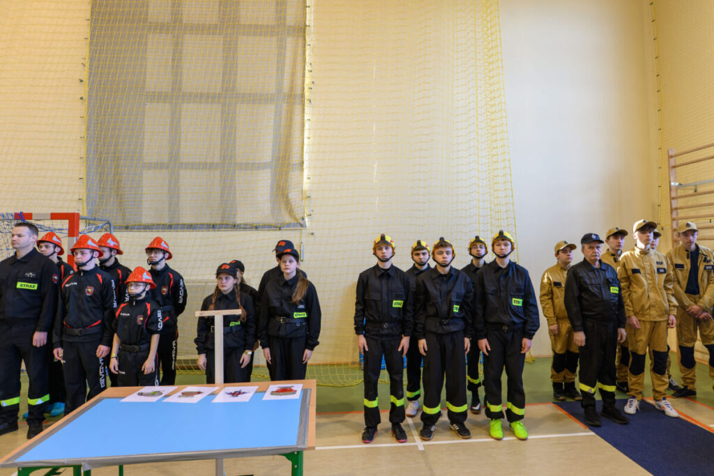 Grupa młodych ludzi w strażackich mundurach stoi w jasnej hali. Mają czerwone i złote hełmy. Na pierwszym planie stół z rysunkami.
