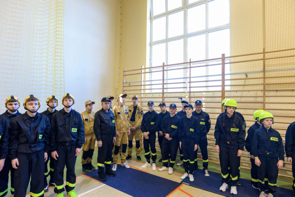 Grupa młodzieży w strojach strażackich stoi w sali gimnastycznej. Noszą granatowe i żółte uniformy z hełmami. W tle są drabinki i duże okno.