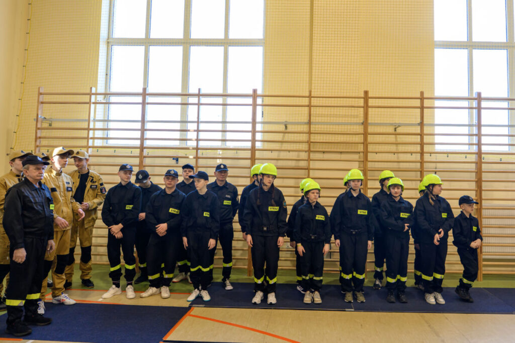 Grupa strażaków w czarnych uniformach i zielonych kaskach stoi w sali gimnastycznej. Za nimi panele i trzy duże okna. Kukatka uniformy mają napisy „STRAŻ”.