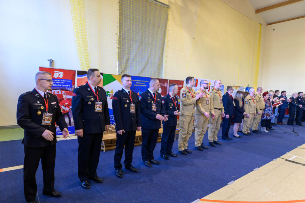 Grupa osób w mundurach stoi w rzędzie na tle kolorowych banerów. Wnętrze ma jasne ściany i niebieską podłogę dywanową. Osoby mają medale na czerwonych wstążkach.