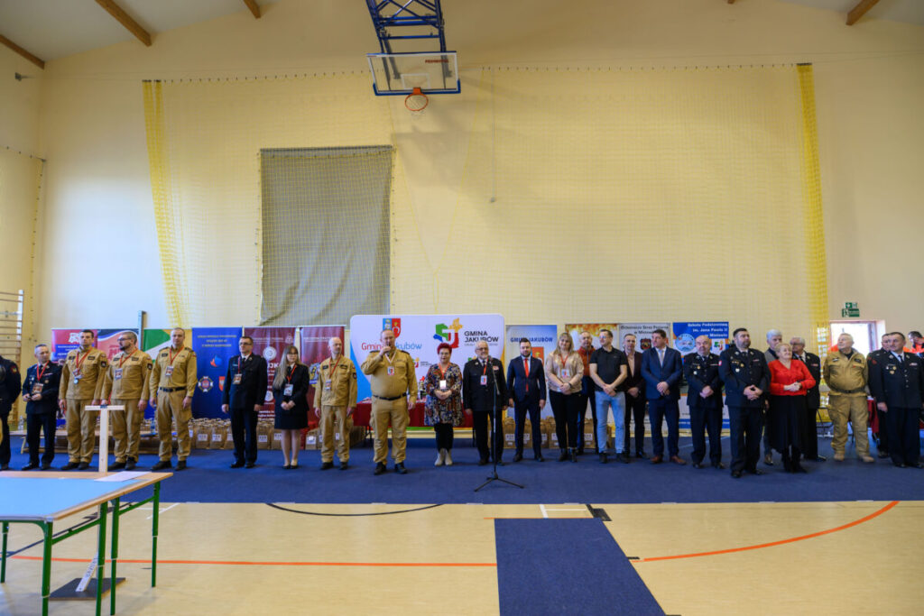 Grupa ludzi w mundurach i ubraniach formalnych stoi w sali gimnastycznej. Tło z banerami promocyjnymi. Podłoga jasna, kosz na suficie.