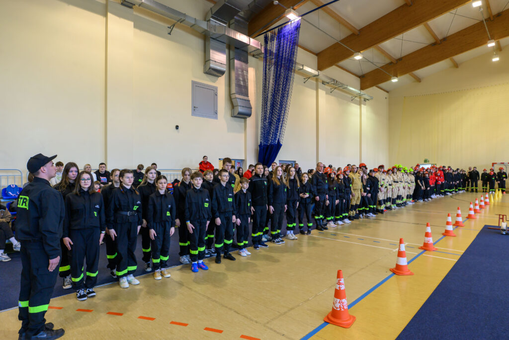 Ludzie w czarnych strojach stoją równo w sali gimnastycznej. Tło ma jasne ściany z niebieskimi zasłonami. Z przodu są pomarańczowe stożki.