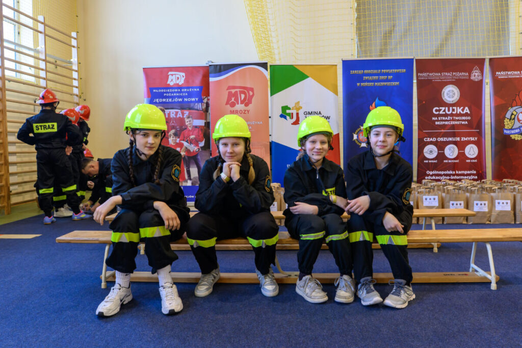 Czwórka dzieci w kaskach i mundurach strażackich siedzi na ławce. Za nimi kolorowe banery i ściana gimnastyczna. Tło jest jasne, z akcentami zieleni i czerwieni.