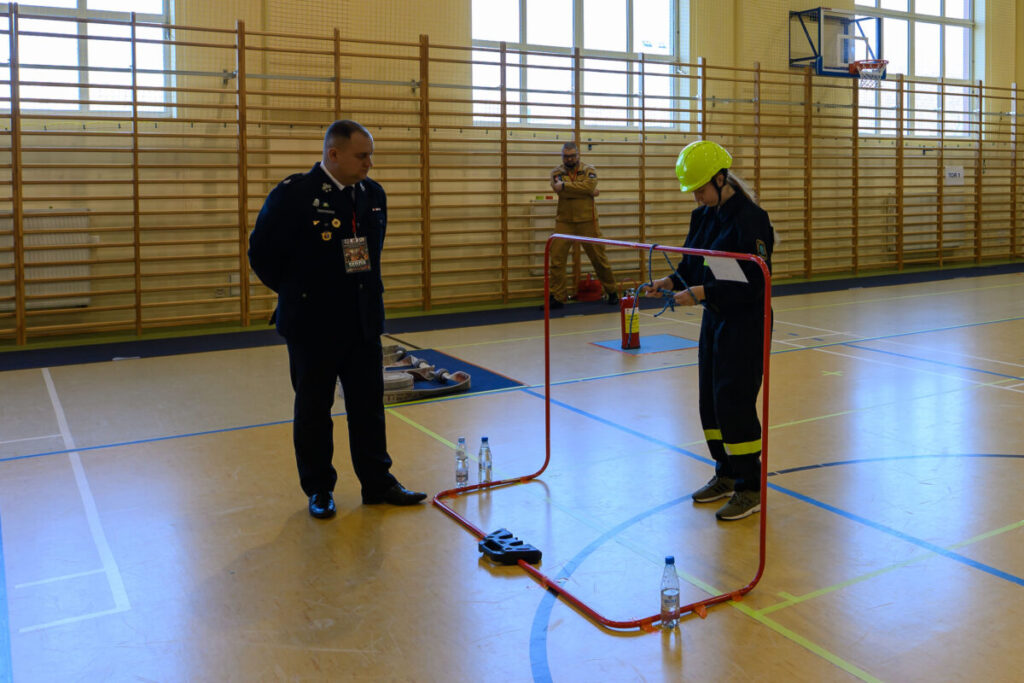 Osoba w mundurze strażackim wykonuje zadanie w sali gimnastycznej. Podłoga jest jasna, w tle widoczne drabinki. Obok osoby stoją butelki z wodą i sprzęt strażacki.