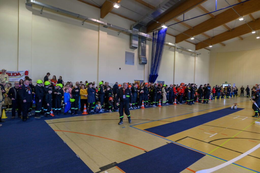 Dorośli i dzieci w kaskach stoją wzdłuż ściany w hali sportowej. Mają na sobie ciemne ubrania z jaskrawymi akcentami. Na podłodze widoczne są niebieskie maty.