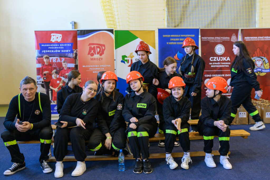 Młodzież ubrana w czarne mundury i pomarańczowe kaski siedzi na ławce. W tle kolorowe banery, podłoga niebieska. Rozmawiają i uśmiechają się.