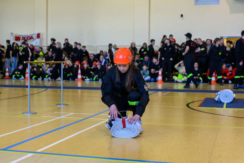 Młoda kobieta w strażackim hełmie zwija wąż na sali gimnastycznej. Tło wypełnia tłum w ciemnych ubraniach. Na podłodze widać niebieskie linie.