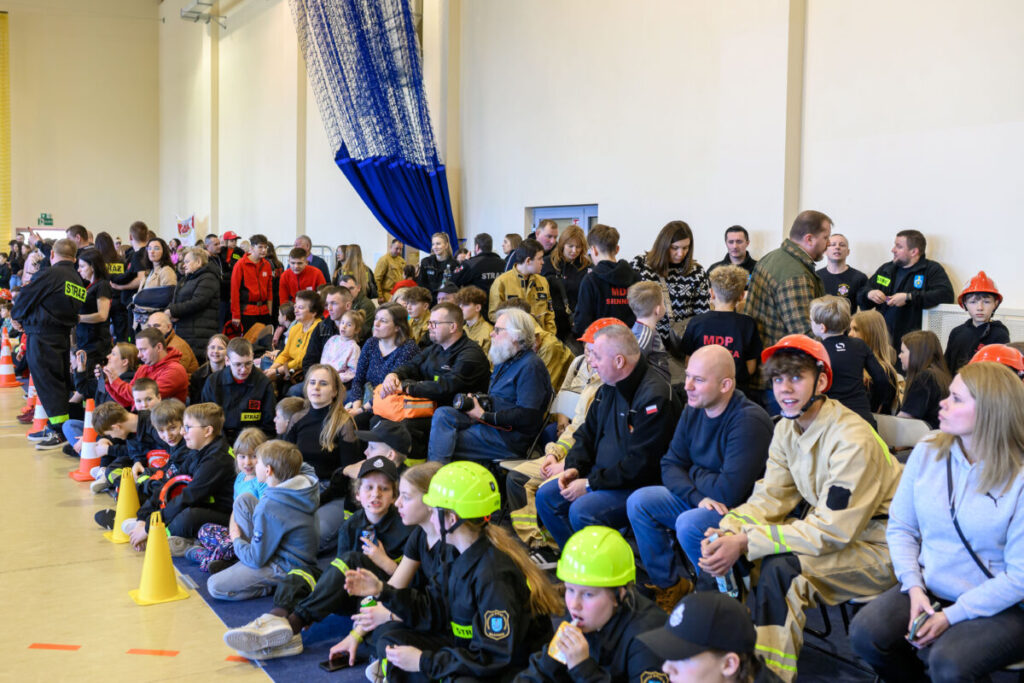 Grupa ludzi w strojach strażackich siedzi na trybunach w jasno oświetlonej hali. Osoby noszą kaski, ubrania w odcieniach czerni i beżu. Widoczne pomarańczowe pachołki na żółtej podłodze.