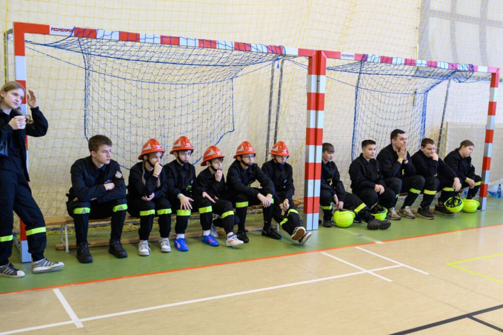 Grupa młodzieży w czarnych uniformach i pomarańczowych kaskach siedzi na ławce w sali gimnastycznej. Tło stanowią bramki z białymi i czerwonymi elementami. Podłoga jest zielono-beżowa z liniami boiskowymi.