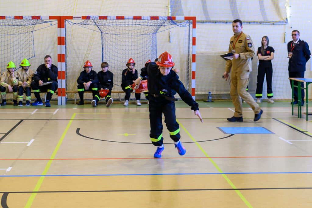 Osoba w stroju strażaka biegnie przez halę sportową. Tło stanowią bramki i obserwujący ludzie w mundurach. Kolory to czerń, czerwony i beżowy.