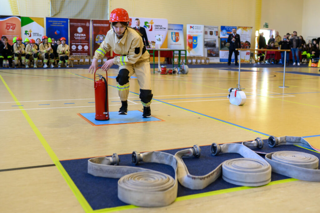 Strażak w czerwonym kasku ćwiczy z gaśnicą na hali sportowej. W tle siedzi grupa strażaków w beżowych mundurach. Na podłodze rozwinięte węże strażackie.