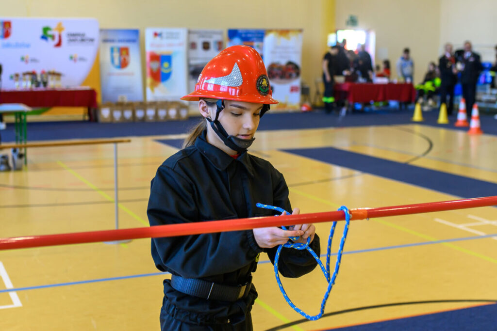 Na zdjęciu młoda osoba w czerwonym hełmie strażackim wiąże niebieską linę. W tle sala gimnastyczna z banerami i stołami. Kolory: czerwony, niebieski i żółty.