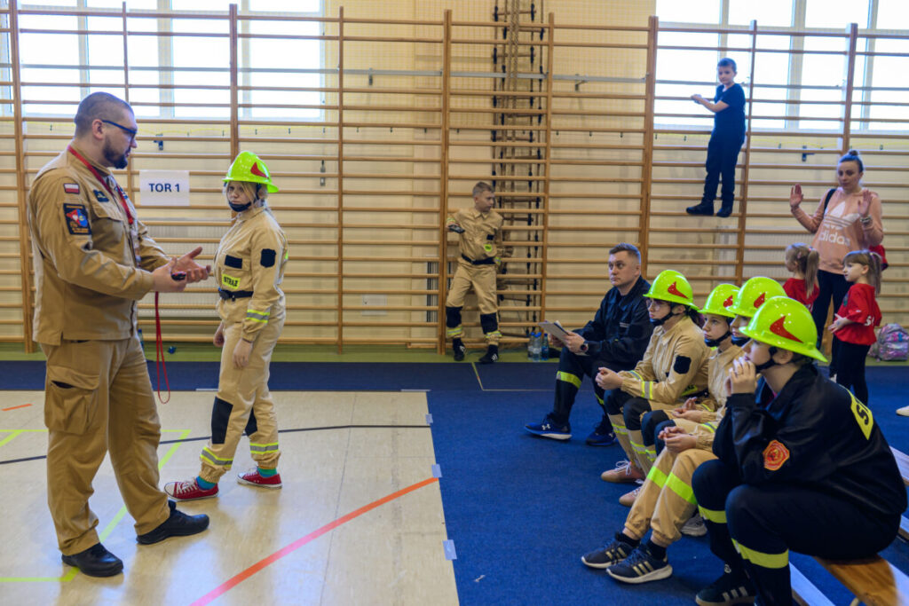 Na zdjęciu w sali gimnastycznej dzieci w strojach strażaków. Żółte hełmy, beżowe stroje. Tło: drewniane drabinki.