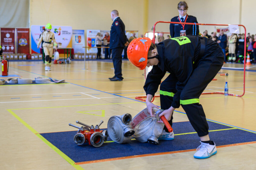 Na zdjęciu młody strażak w kasku i stroju układa wąż strażacki na hali. W tle tablice informacyjne i ludzie. Dominują kolory czerwony, żółty i czarny.